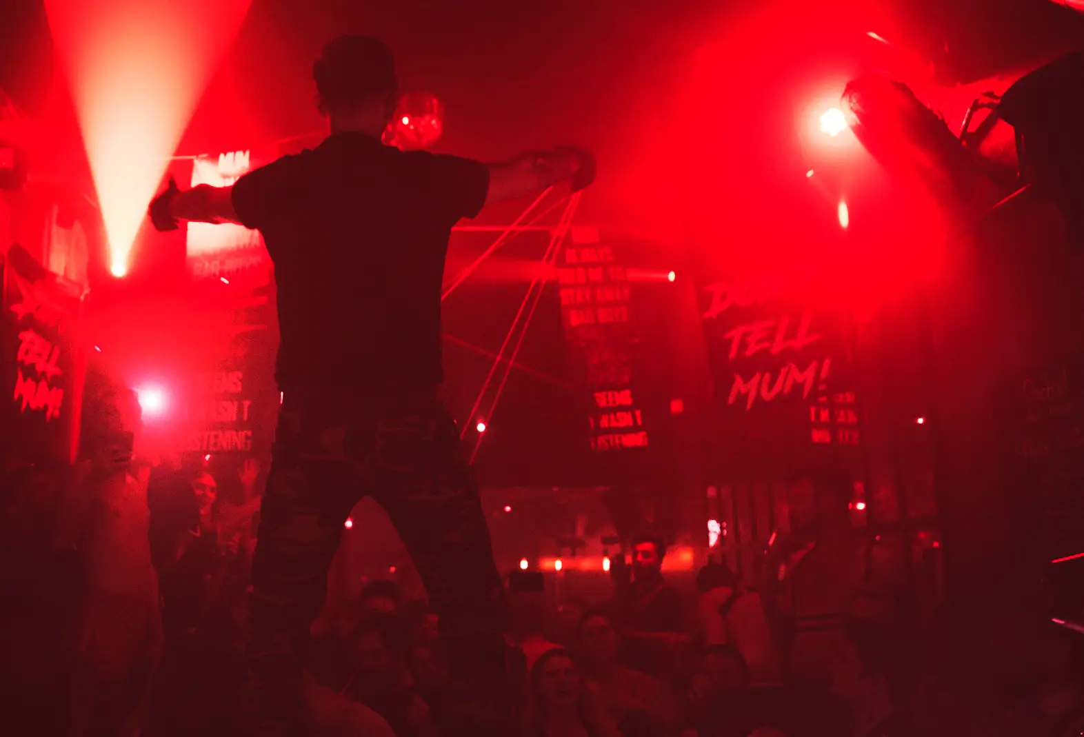 Silhouette sur estrade bras ouverts sous lumière rouge, foule et bannières Don't Tell Mum lors d’une soirée fetish Paris